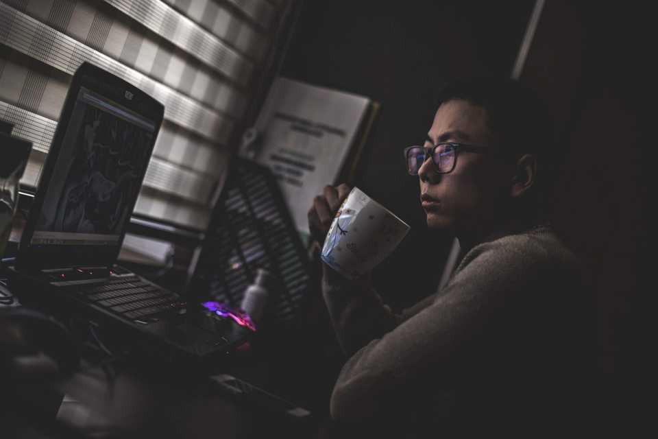 Desenvolvedor, dentro do que parece ser seu quarto, tomando algo de uma xícara, olhando atentamente para a tela do notebook.