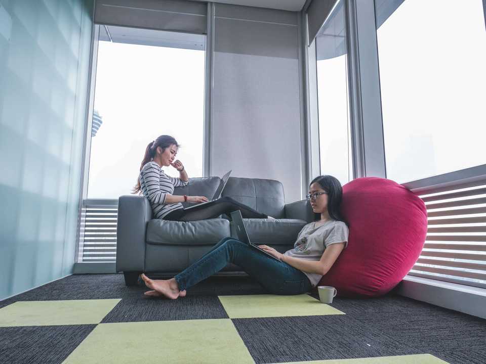 Na imagem, duas garotas estão concentradas, cada uma com o seu note. Uma está sentada no sofá e a outra no chão, num puff.