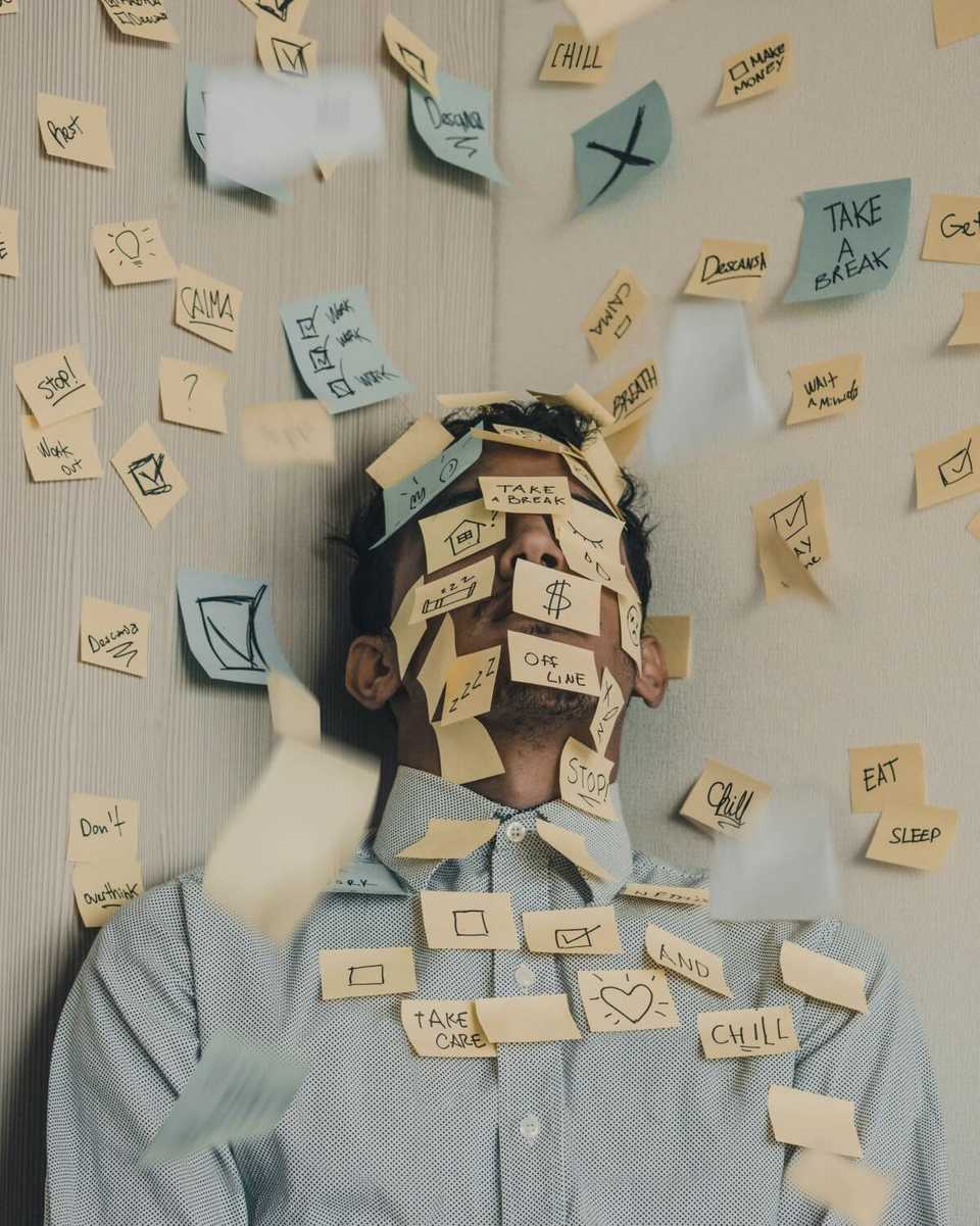 Imagem de um homem com camisa social, aparentemente em um escritório, e coberto de post-its (inclusive no rosto).