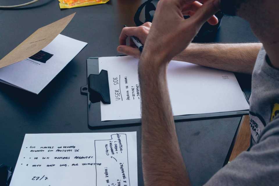 Homem concentrado, debruçado sobre uma prancheta e tomando notas sobre experiência do usuário. Na mesa, há mais papéis com outras anotações.